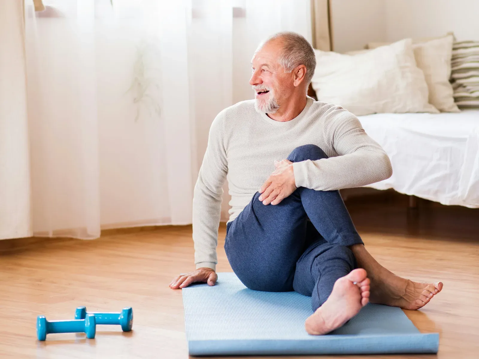 A man in his seventies sitting on a mat at home, hugging one knee and laughing, light dumbbells beside him