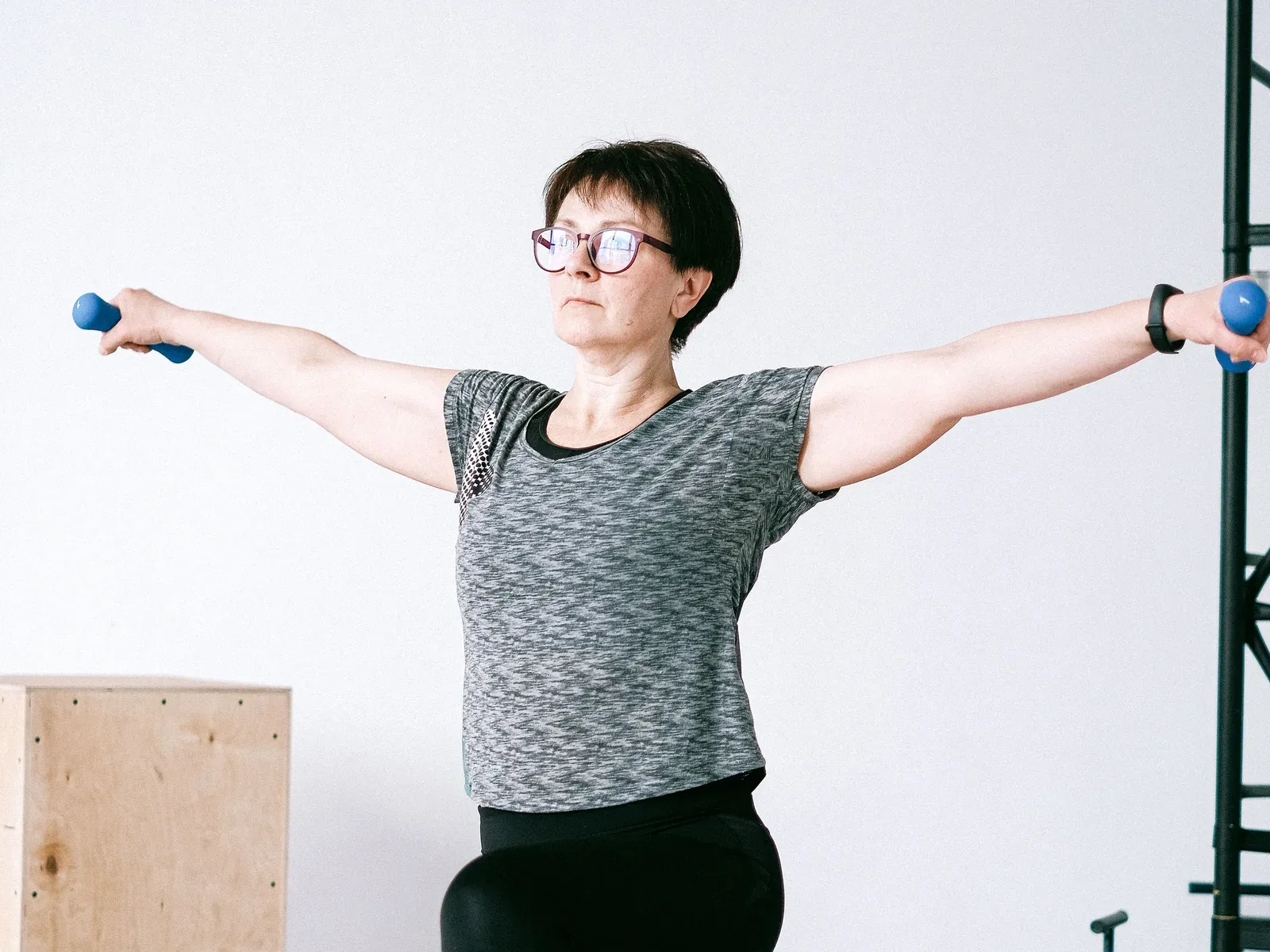 A woman in her fifties in a kneeling lunge, arms out to the sides holding light dumbbells